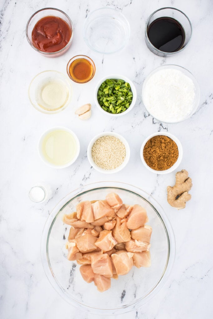 Overhead shot of raw chicken pieces in a glass bowl surrounded by small bowls of sesame chicken ingredients like ketchup, soy sauce, cornstarch, garlic, ginger, green onions, sesame seeds, and seasonings on a marble surface.