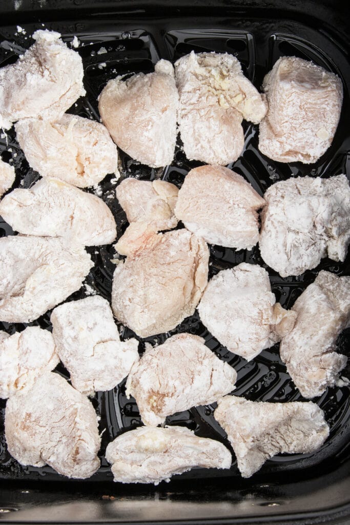 Chicken pieces coated in cornstarch arranged in a single layer inside an air fryer basket, sprayed lightly with oil.
