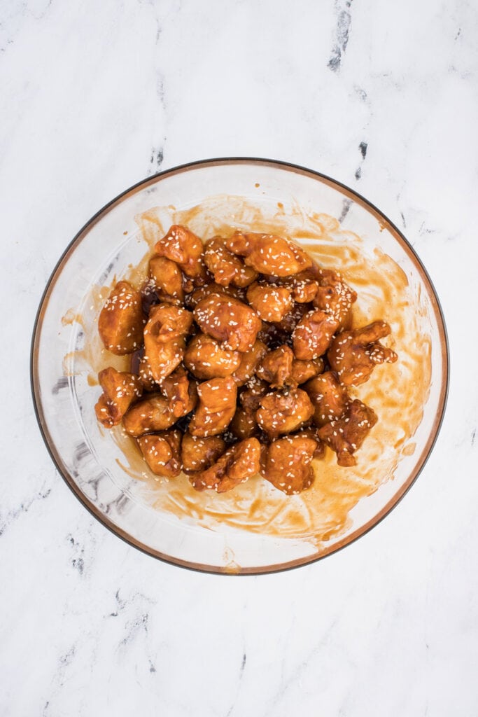 Finished sesame chicken in a glass bowl coated with sauce and sprinkled with sesame seeds, ready to serve.