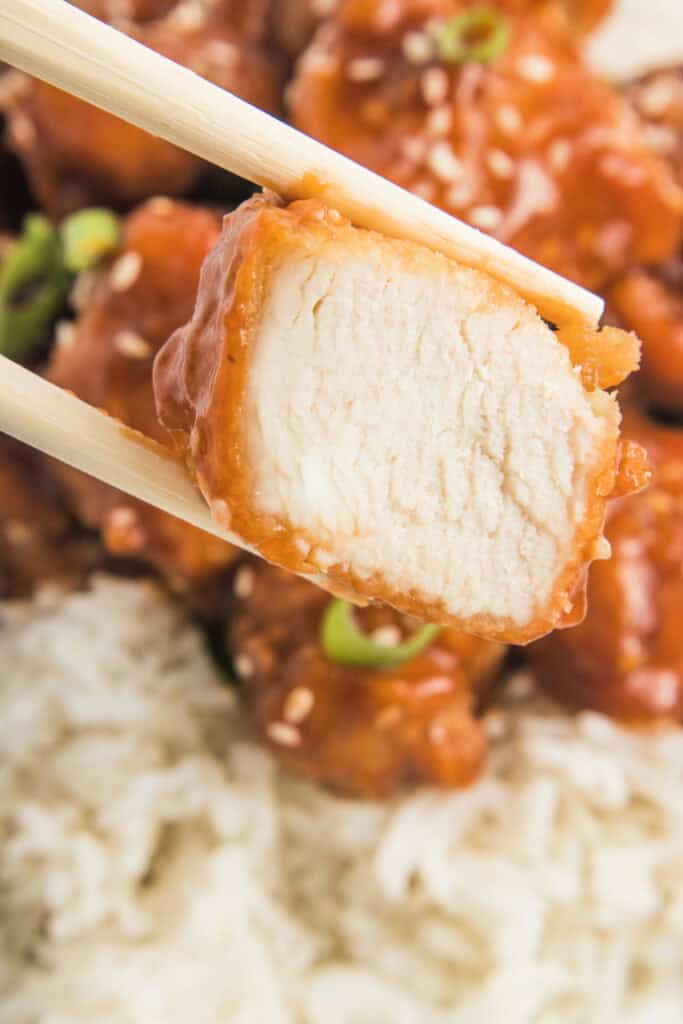 Chopsticks holding a piece of sesame chicken showing the juicy cooked inside, with rice and more chicken blurred in the background.