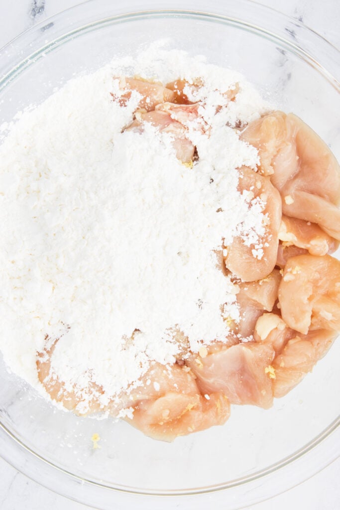 Chicken chunks in a glass bowl covered with a generous layer of white cornstarch before tossing.
