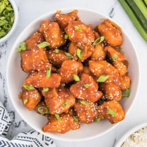 Square crop of a bowl filled with saucy sesame chicken, garnished with sesame seeds and green onions, styled with rice and fresh garnishes on the side.