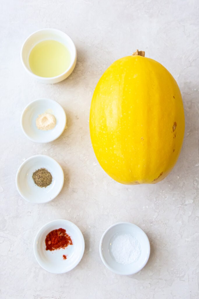 Whole spaghetti squash with small bowls of oil, salt, pepper, paprika, garlic powder, and Italian seasoning on a light background.