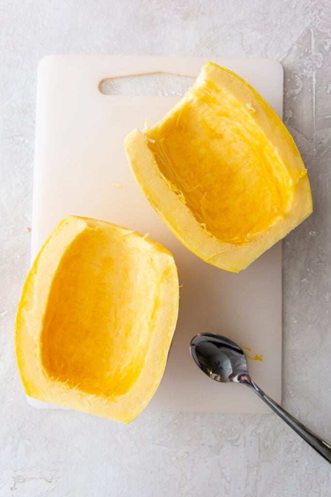Two spaghetti squash halves with seeds removed, cleaned out and ready for cooking, on a cutting board with a spoon.