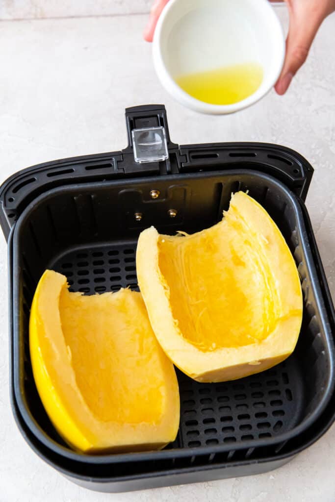 Hand holding a small bowl of oil over two spaghetti squash halves in the air fryer basket.