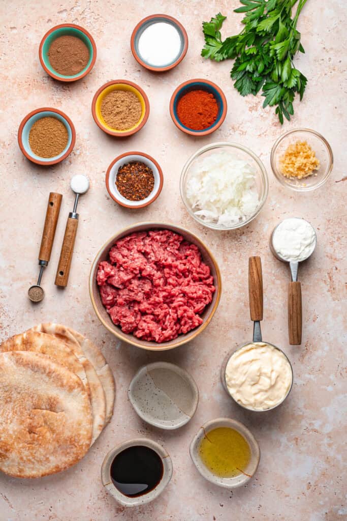 Overhead shot of arayes ingredients including ground beef, ground lamb, spices in small bowls, parsley, pita bread, mayonnaise, yogurt, olive oil, and pomegranate molasses on a light surface.