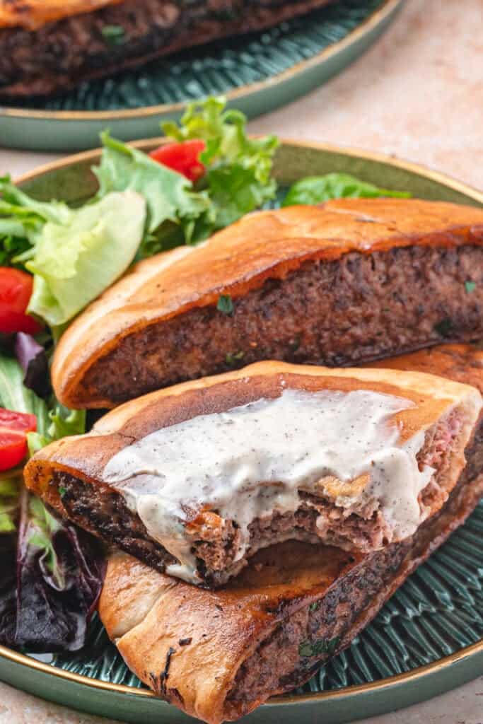 Arayes served on a green plate with salad, one piece topped with creamy shwarma white sauce and a bite taken to show texture.