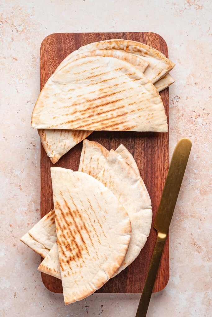 Stack of halved pita bread pieces arranged on a wooden cutting board with a gold knife, ready to be stuffed for arayes.