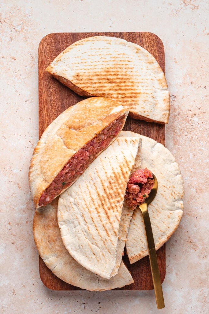 Pita halves stuffed with raw spiced meat mixture arranged on a wooden board with a gold spoon tucked inside one pita showing the filling.