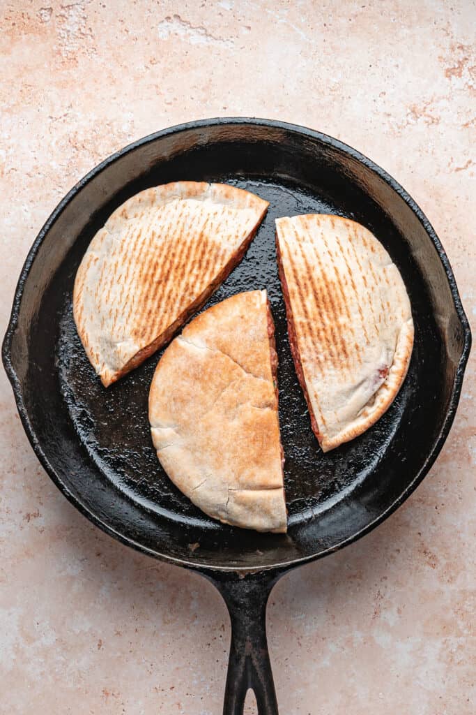 Three raw stuffed pitas cooking in a cast iron skillet, open side down, beginning to sear for arayes.