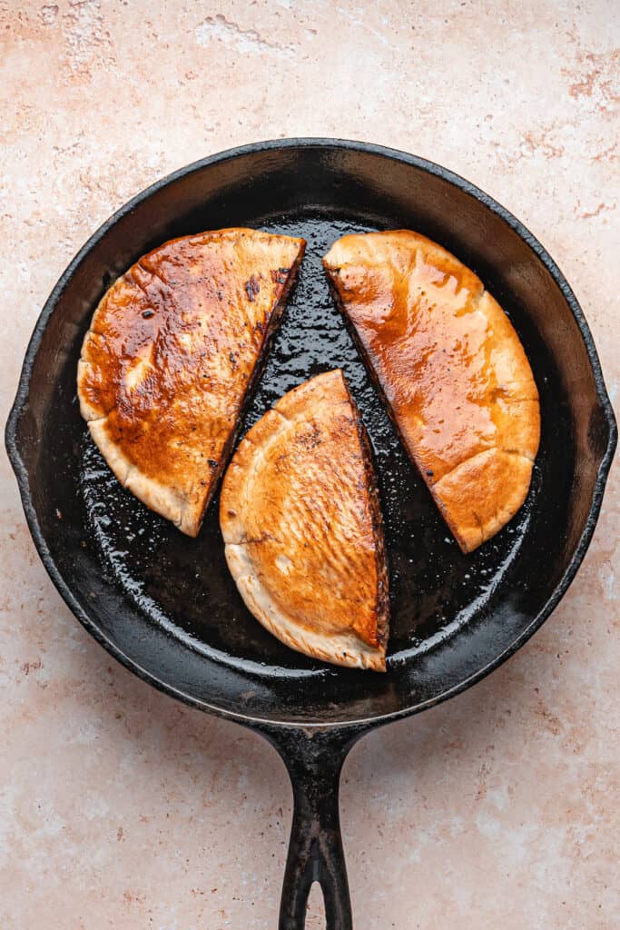 Three stuffed pitas flipped in a cast iron skillet, golden brown and crisping on the surface as they cook for arayes.