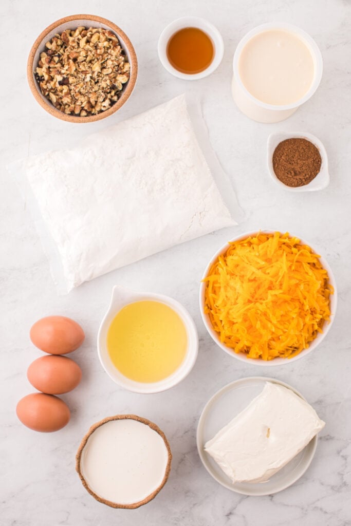 Overhead view of butternut squash cake ingredients including walnuts, eggs, grated squash, cream, oil, cream cheese, cake mix, maple syrup, and spices arranged on a white surface.