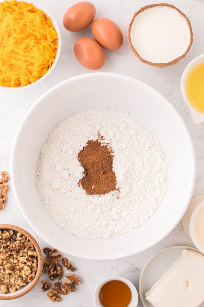 Mixing bowl with white cake mix and pumpkin pie spice surrounded by eggs, grated squash, cream, oil, walnuts, and cream cheese.