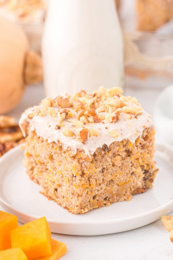 Slice of butternut squash cake on a white plate, topped with frosting and walnuts, with milk bottle in background.