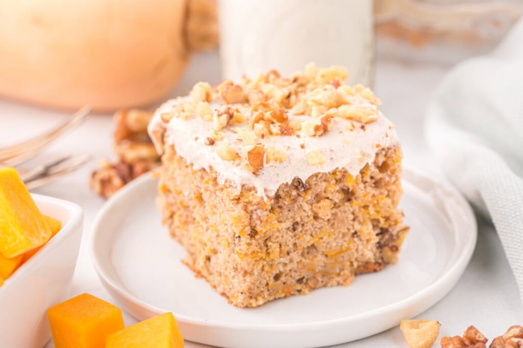 Wide view of a slice of butternut squash cake on a plate with walnuts and squash pieces around it.