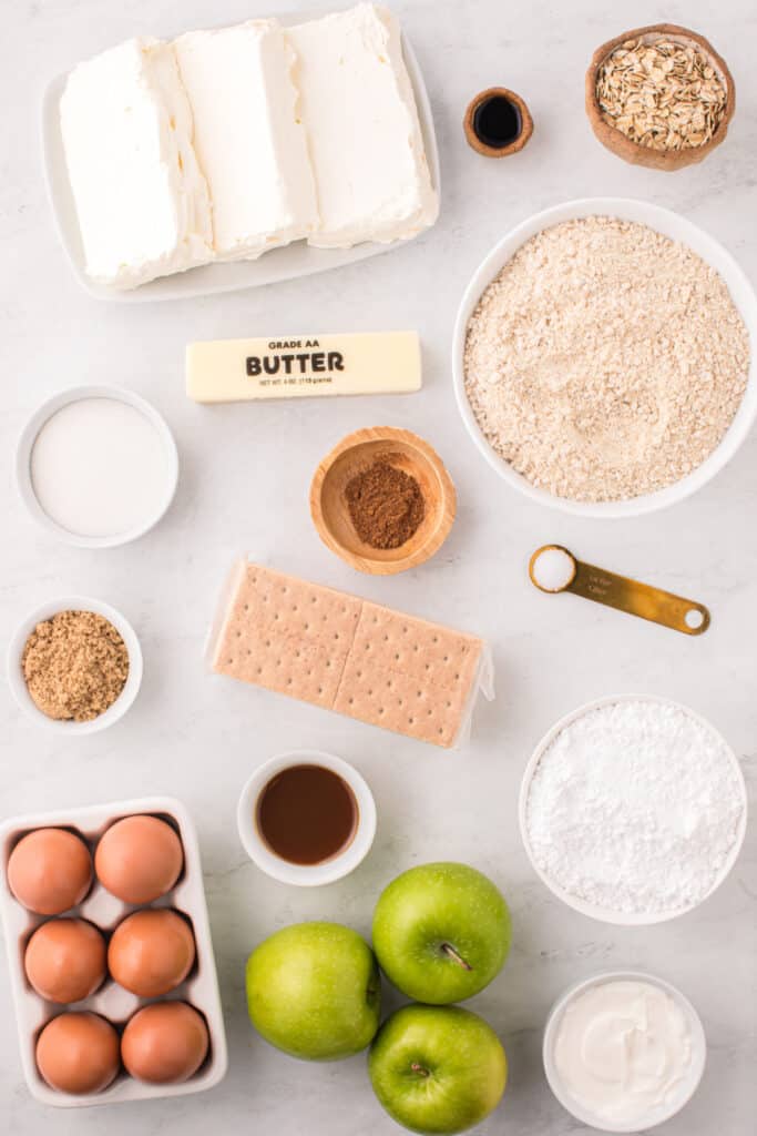 Overhead view of cheesecake bar ingredients including cream cheese, butter, sugar, graham crackers, oats, apples, eggs, and spices.