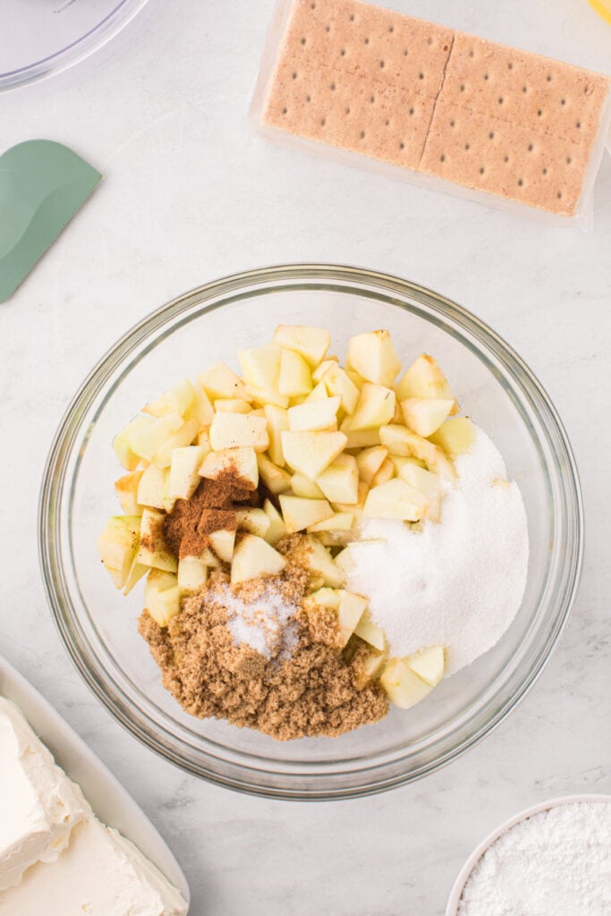 Bowl of chopped apples topped with sugar, brown sugar, salt, and pumpkin pie spice ready to be mixed.