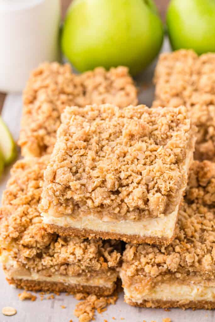 Stack of cheesecake bars with visible apple filling and crumb topping, apples in background.