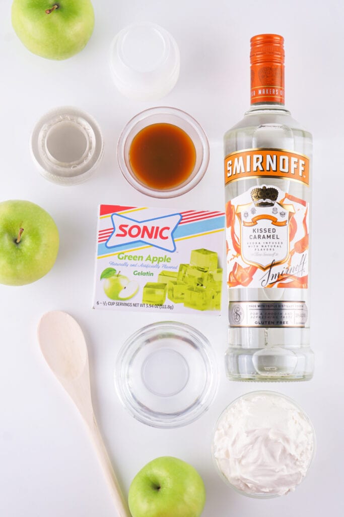 Ingredients for caramel apple jello shots including green apples, Sonic green apple gelatin, caramel vodka, caramel sauce, water, and whipped topping.