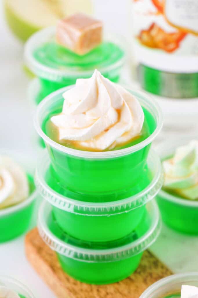 Stacked green caramel apple jello shots in plastic cups, topped with whipped cream and caramel drizzle.