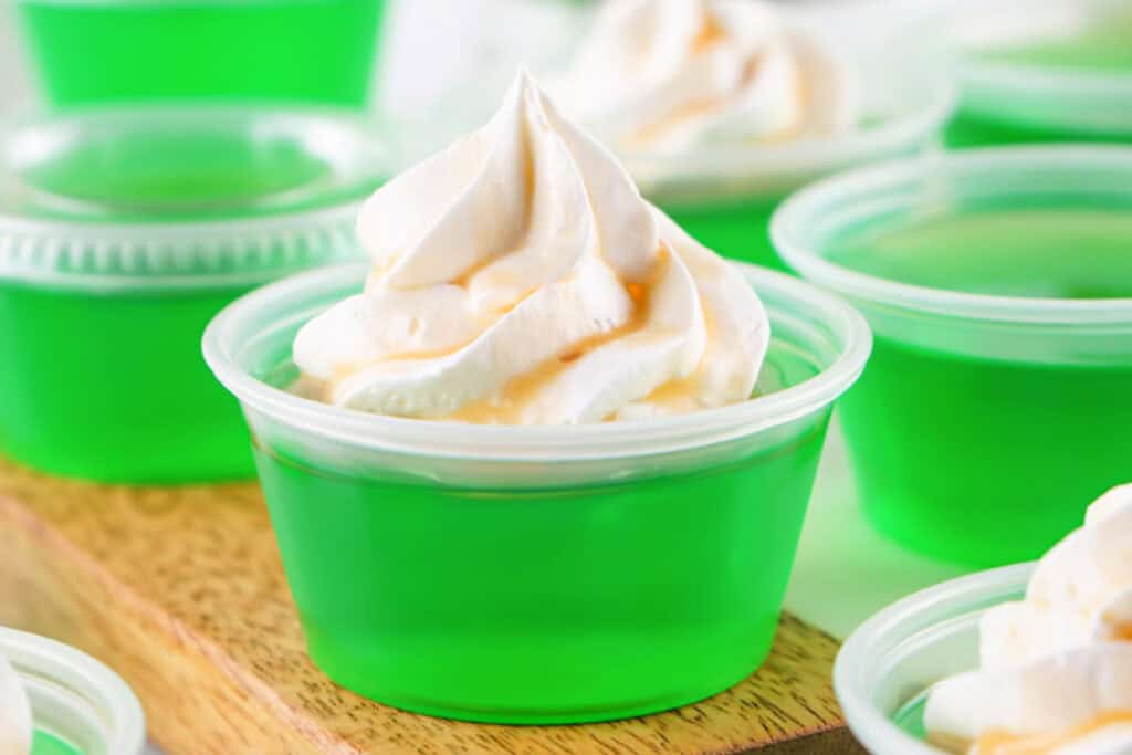Close-up of a single green jello shot topped with whipped cream and caramel drizzle, surrounded by other shots.