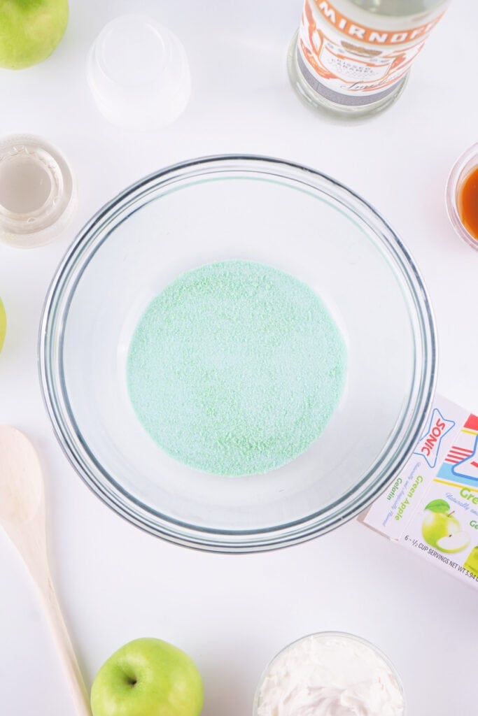Green apple jello powder poured into a clear mixing bowl, surrounded by ingredients for jello shots.