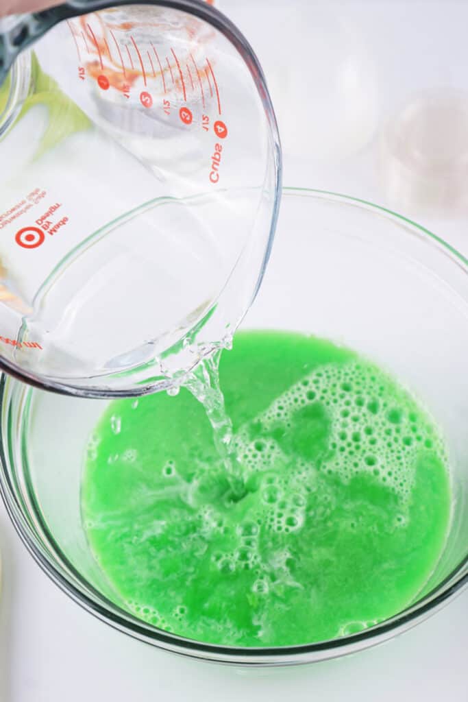 Measuring cup of boiling water being poured into a bowl of green apple jello powder, creating a bright green mixture.