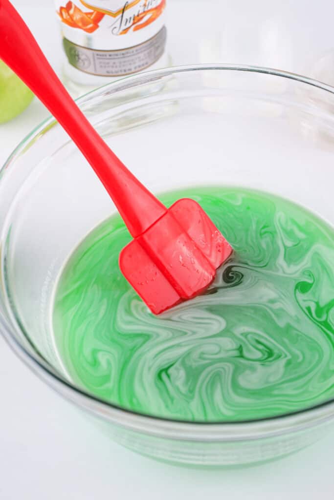 Red spatula stirring the dissolved green apple jello mixture in a clear glass bowl.