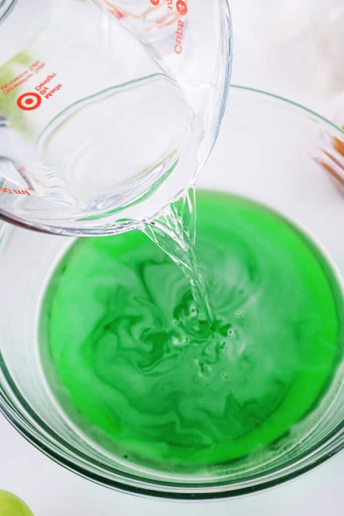 Measuring cup pouring vodka into the prepared green apple jello mixture.