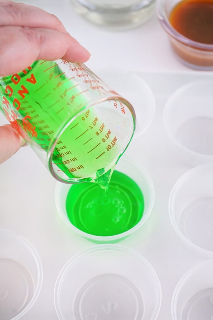 Green jello mixture being poured into small clear plastic shot cups from a measuring cup.
