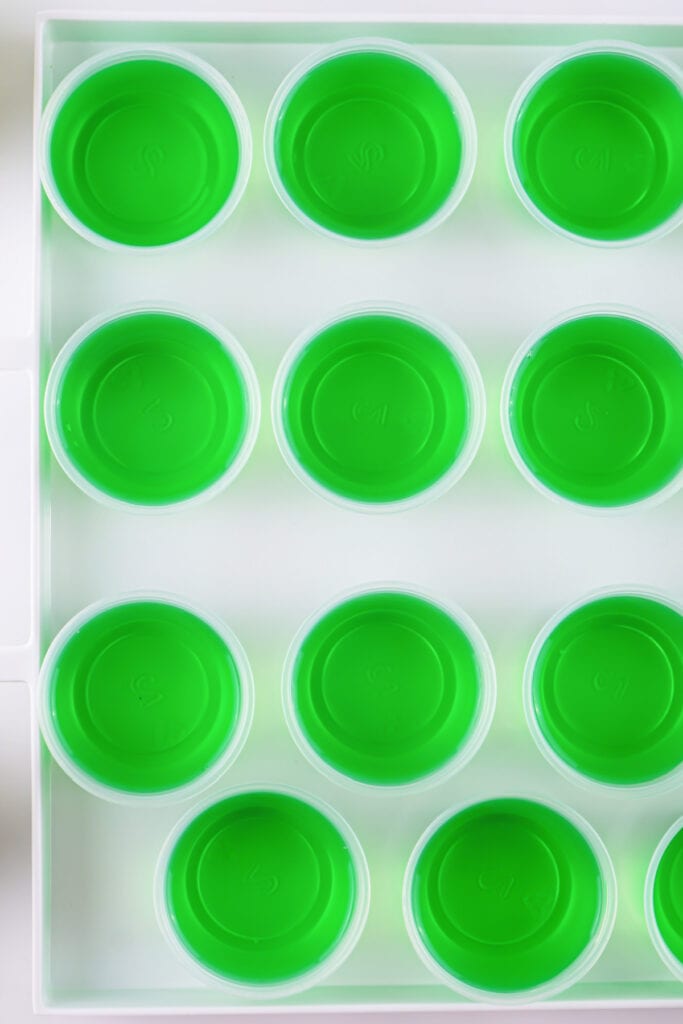 Tray of small plastic cups filled with bright green jello setting in neat rows.