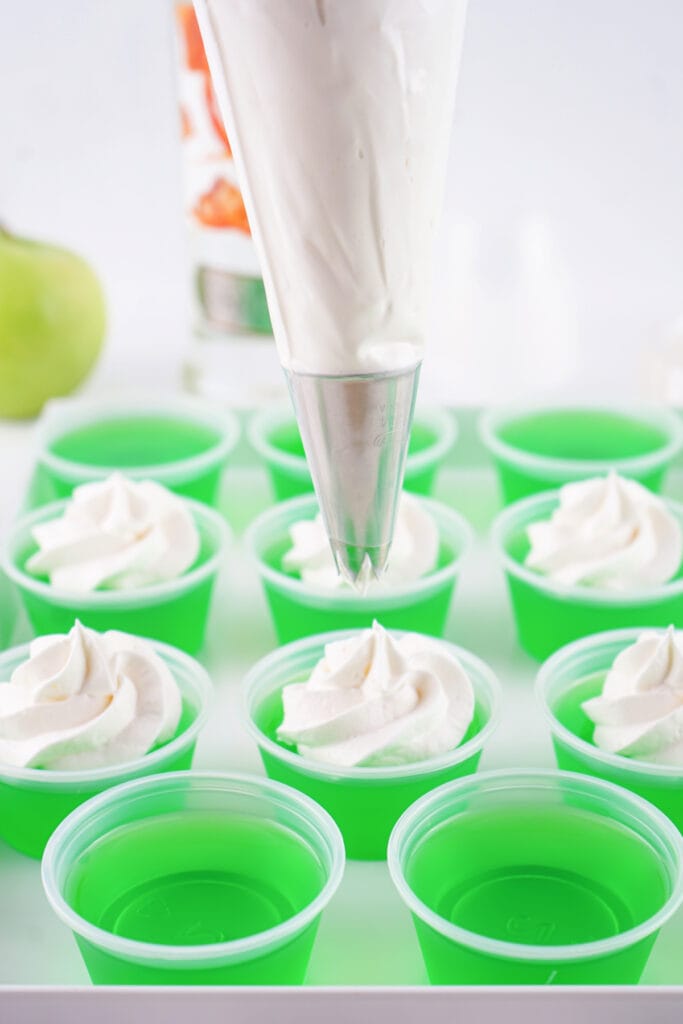 Piping bag fitted with a star tip adding whipped topping to the set green jello shots.