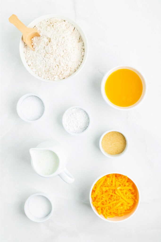 Overhead view of measured ingredients for Cheddar Butter Swim Biscuits including flour, cheddar, butter, milk, salt, and seasonings.