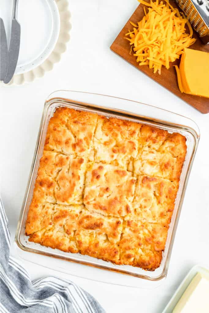 Baked Cheddar Butter Swim Biscuits in a glass pan, golden brown and cut into squares, with shredded cheddar on the side.