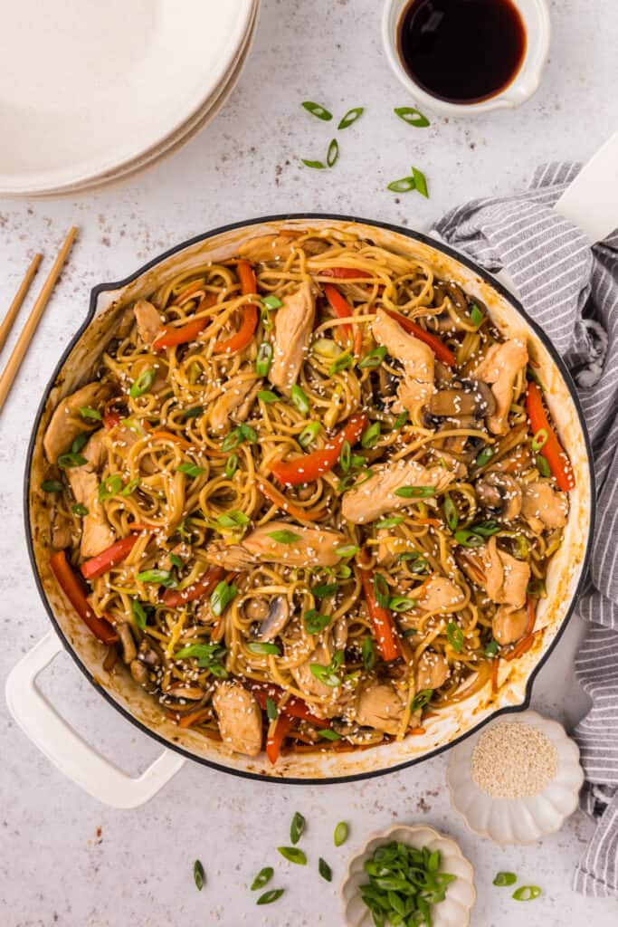 Overhead shot of Chicken Lo Mein in a white skillet, garnished with sesame seeds and green onions with plates and chopsticks nearby