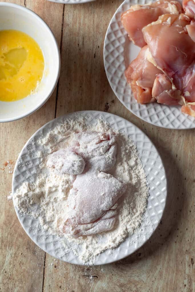 Chicken thighs dredged in flour on a white plate, with eggs and raw chicken ready for the breading process.