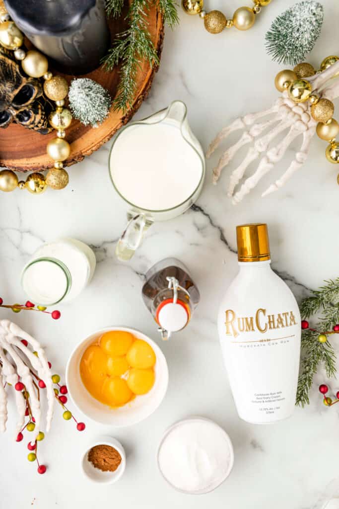 Overhead photo of cocktail ingredients including RumChata, eggs, sugar, milk, cream, pumpkin spice, and vanilla surrounded by festive Halloween and Christmas decorations.
