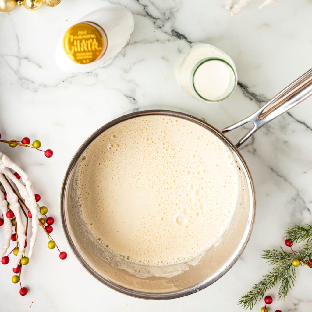 Saucepan filled with smooth, foamy eggnog base after cooking, resting on marble with festive decor nearby.