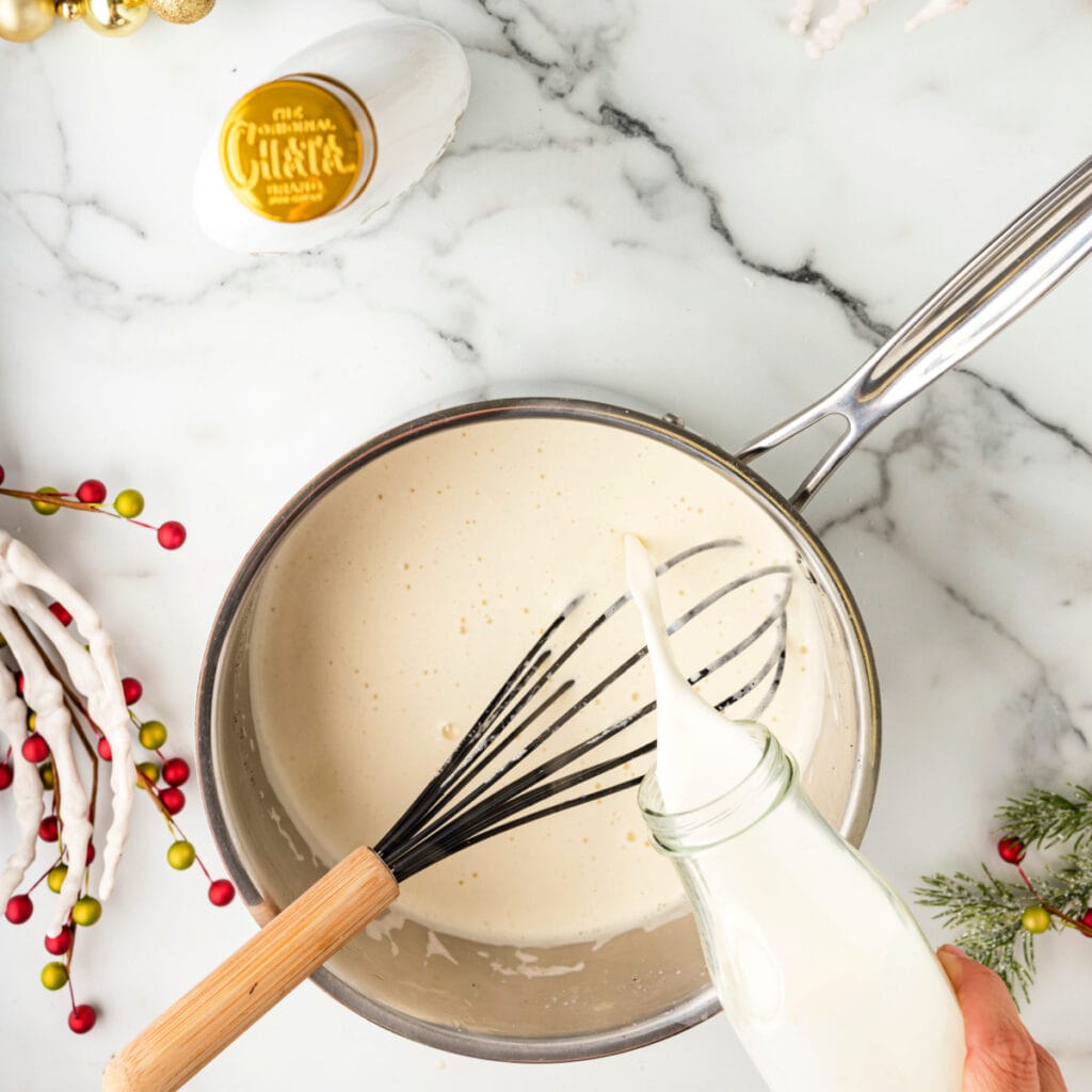 Hand pouring cream into a saucepan of warm eggnog mixture while whisking.