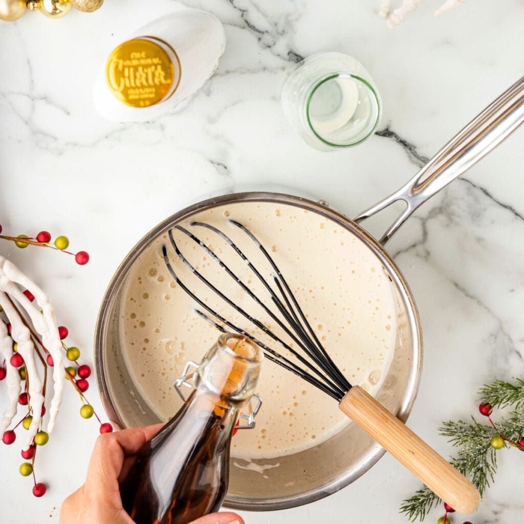 Hand pouring vanilla extract into a saucepan of cooked eggnog mixture with a whisk resting inside.
