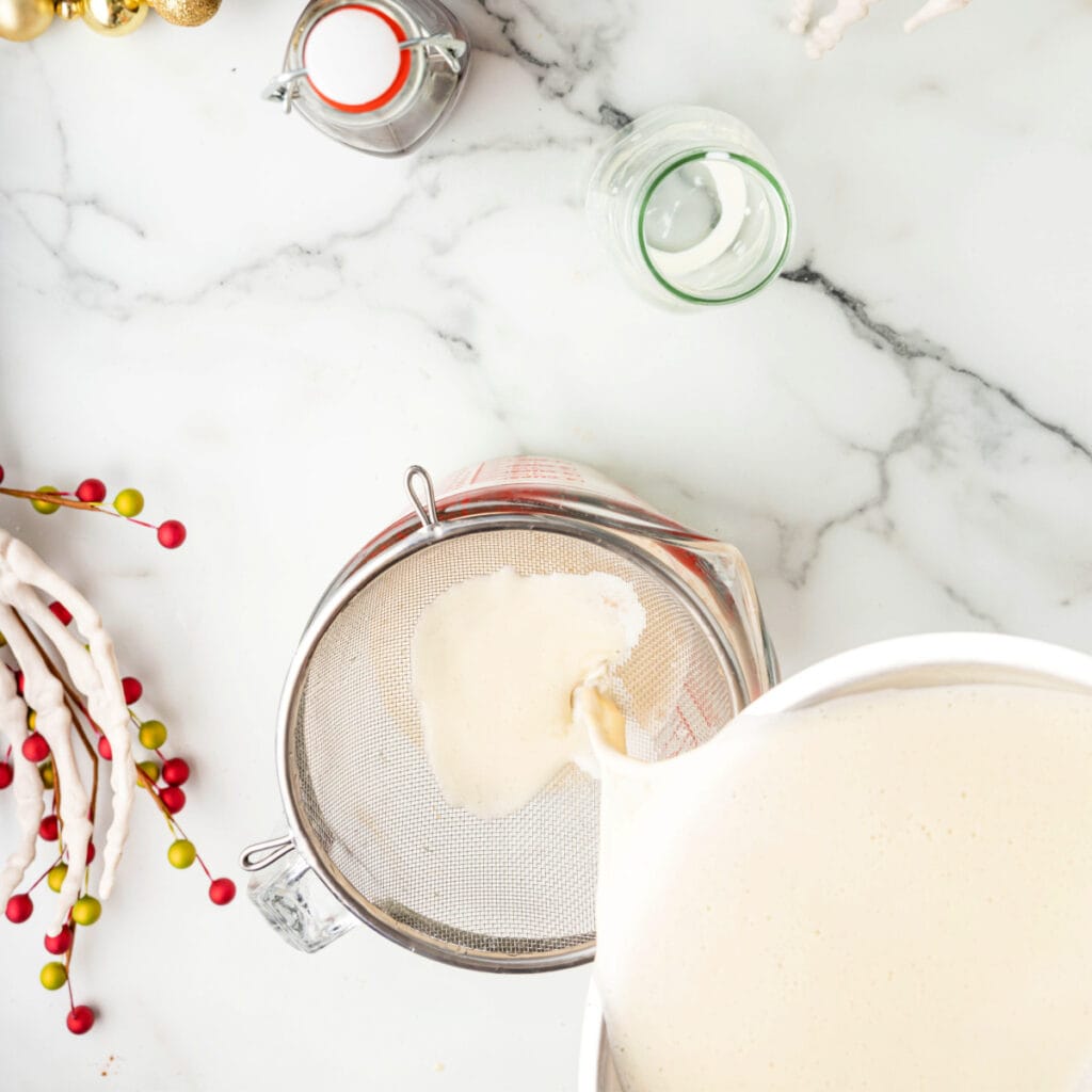 Straining eggnog base through a fine mesh strainer into a glass measuring cup.