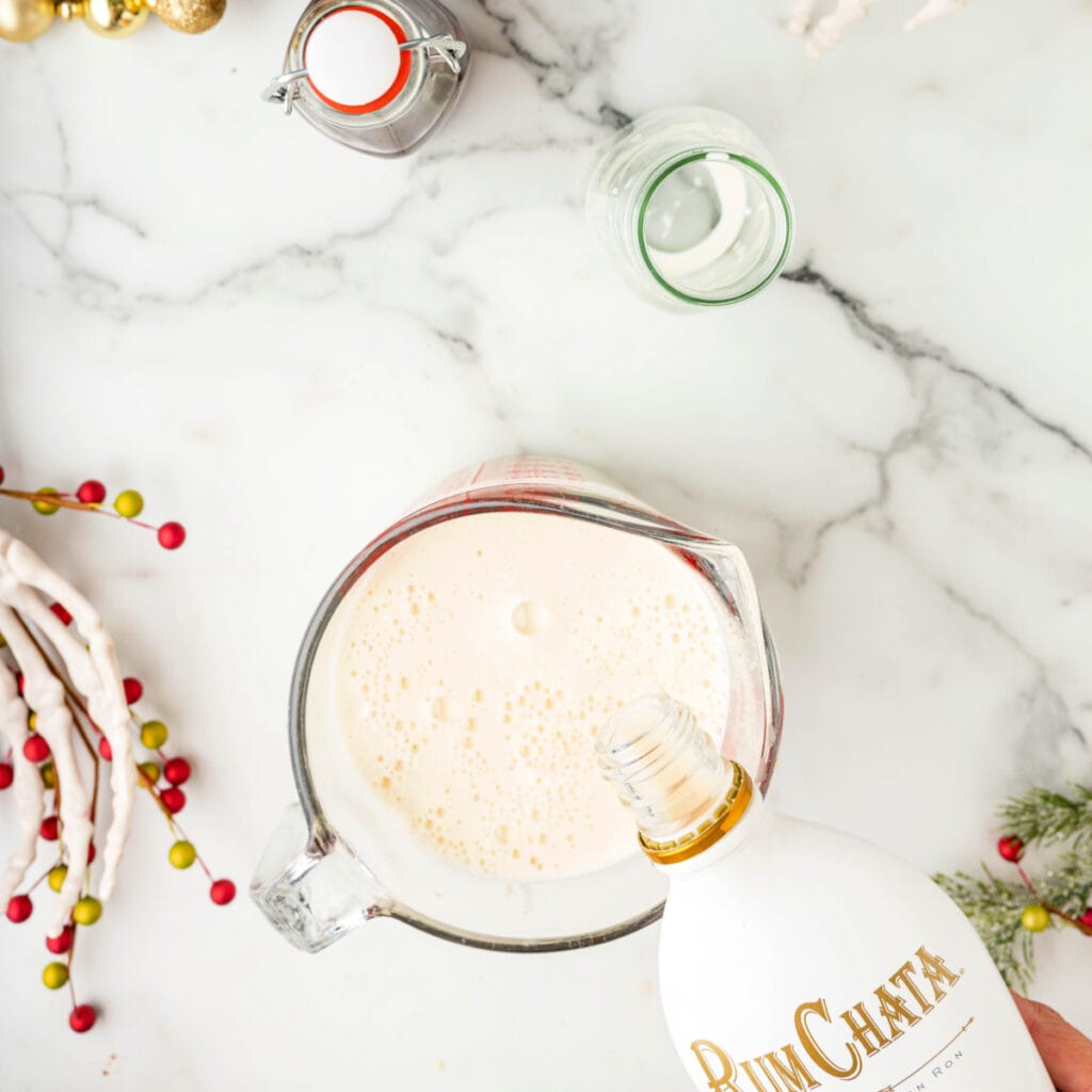 Hand pouring RumChata into a glass measuring cup filled with cooled eggnog base.