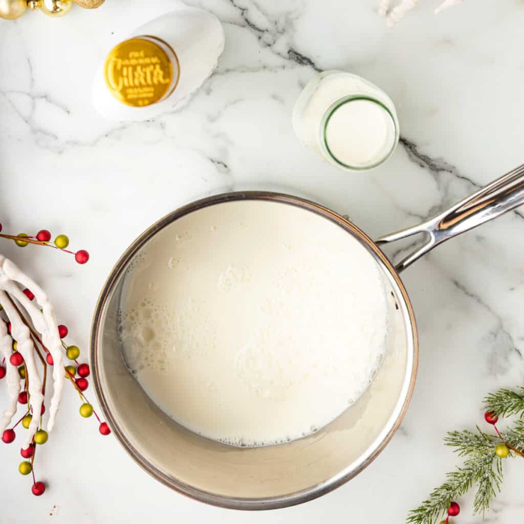 Saucepan filled with milk heating on the stove, RumChata and cream jar nearby on a marble surface with holiday decor.