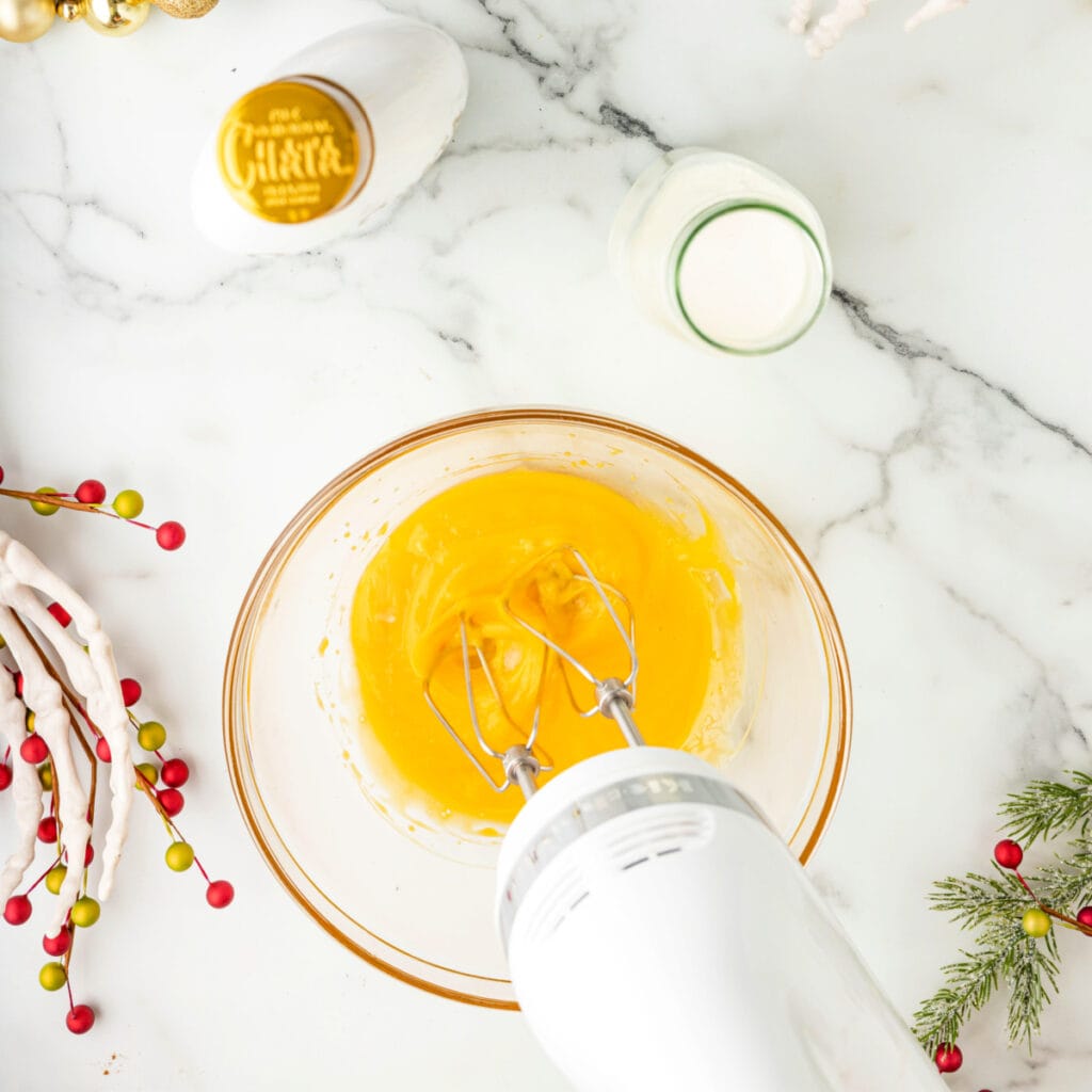 Electric mixer blending bright yellow egg yolks in a glass bowl on a decorated marble counter.