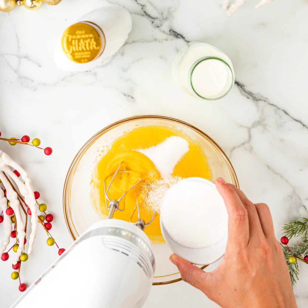 Hand pouring sugar into beaten egg yolks while using an electric mixer in a glass bowl.