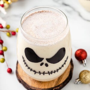 Close-up of a Jack Skellington Cocktail served in a glass decorated with Jack’s face, sitting on a wooden coaster with holiday ornaments nearby.
