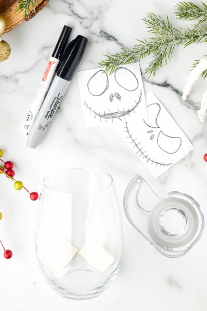 Supplies laid out on a white marble surface including sharpies, tape, and Jack Skellington face templates next to a clear glass.