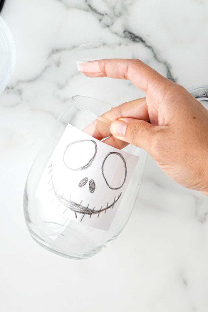 A hand placing the Jack Skellington face template inside a clear glass and securing it with tape.
