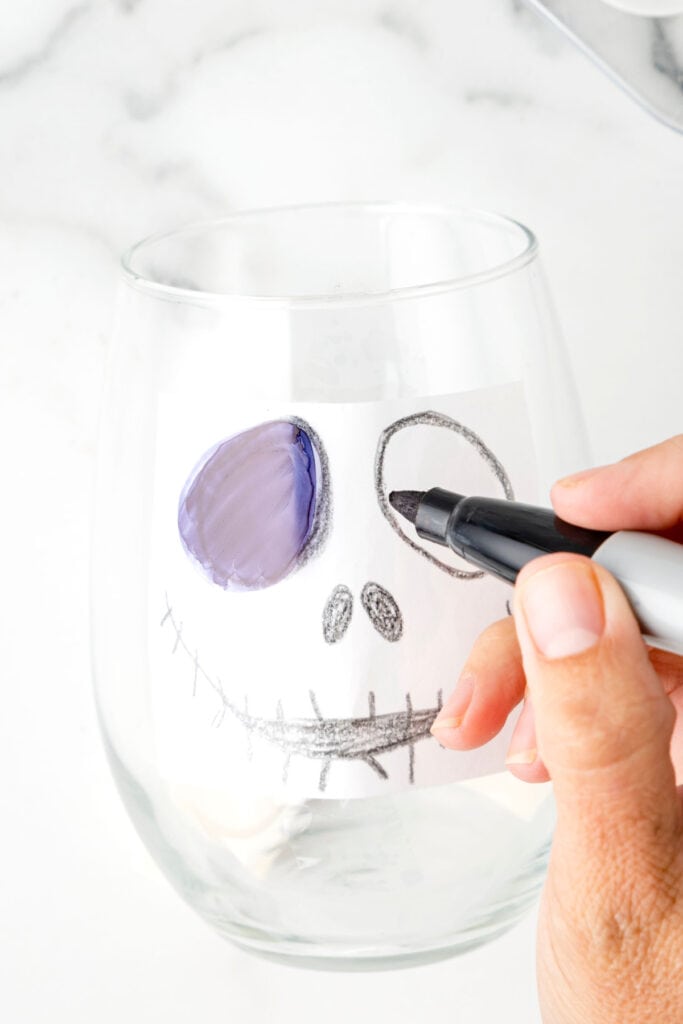 A close-up of tracing the Jack Skellington face template on the glass with a black sharpie, beginning to fill in the eyes.