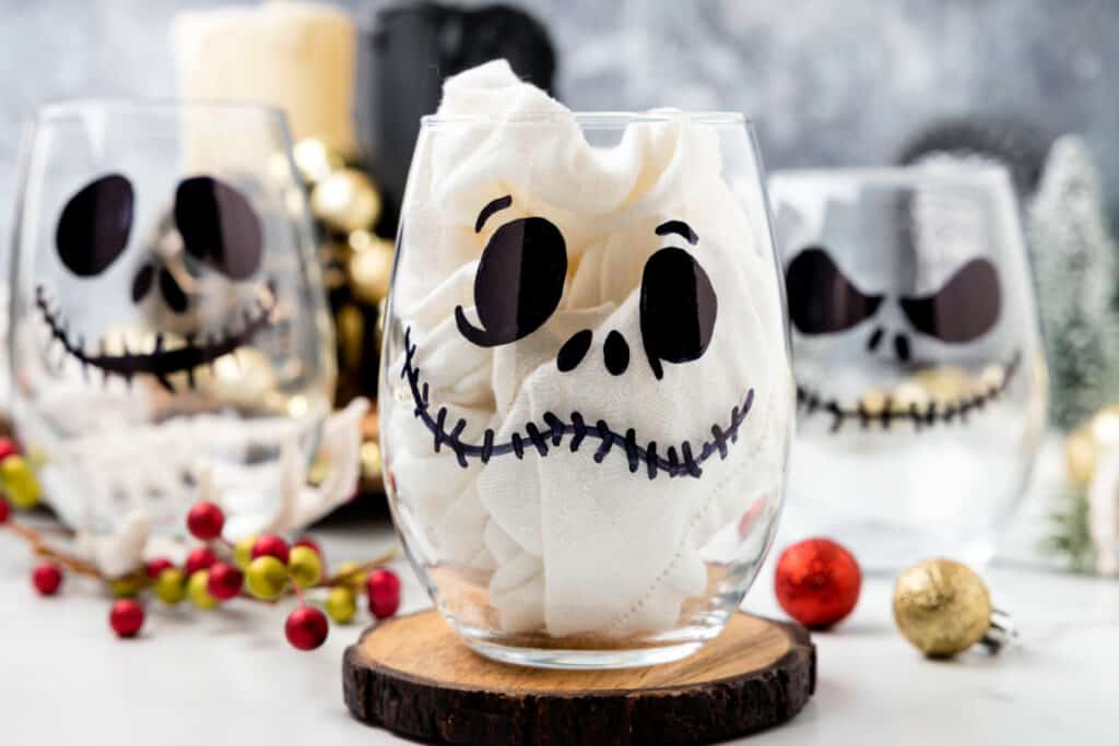 Three finished Jack Skellington glasses on a holiday table setting with ornaments and greenery.